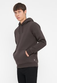 Brown cotton hoodie with a front pocket, drawstring hood, and ribbed cuffs, paired with black pants, set against a plain background.