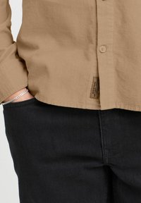 Camisa de pana beige con frente abotonado, mangas largas y etiqueta lateral, combinada con pantalones de mezclilla negros. Tejido texturizado visible.