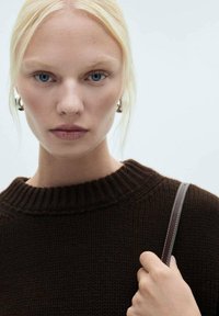 Brauner Strickpullover mit einem gerippten Halsausschnitt, getragen von einer Person mit glattem, blonden Haar. Ein dünner Riemen wird in der rechten Hand gehalten.