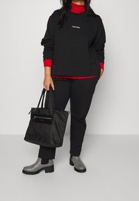 Svart sweatshirt med röda accenter, digitalt logo. Matchande svarta byxor, grå ankelskor med tjocka sulor, och en svart tote med dragkedjedetalj.