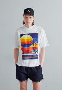 White short-sleeve T-shirt featuring a vibrant graphic of a sunset with palm trees. Paired with dark denim shorts, creating a casual look.