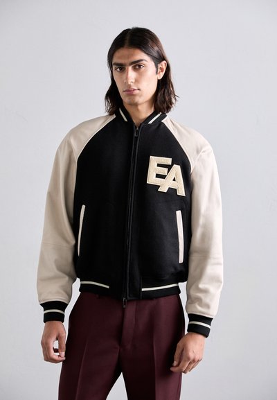 Joven con cabello oscuro de longitud hasta los hombros que lleva una chaqueta de béisbol negra y beige con las letras "EA" y pantalones burdeos, sobre un fondo liso.