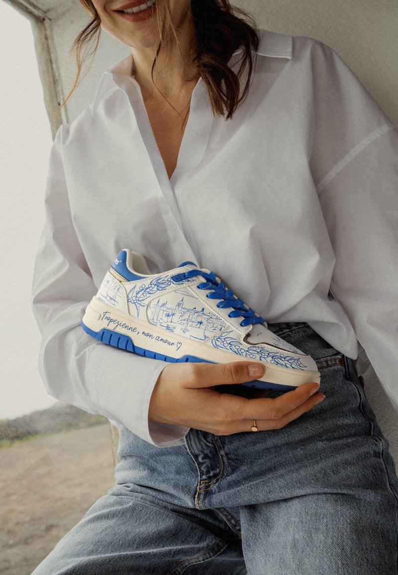 Zapatilla blanca con detalles azules y cordones. Presenta una ilustración detallada en azul y texto en el lateral. Material superior ligeramente texturizado.