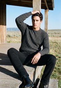 Jeune homme en pull sombre et pantalon assis sur un porche en bois, reposant la main sur la tête, avec un paysage herbeux ouvert et un ciel bleu derrière.