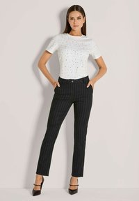 Vrouw die staat met haar handen in haar zakken, gekleed in een wit T-shirt met korte mouwen met kleine stippen en zwart-gestreepte broek met zwarte hakken.