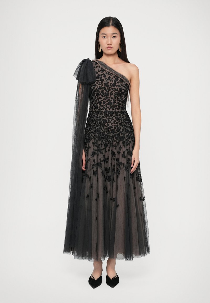 Femeie îmbrăcată într-o rochie din tull neagră cu un umăr, cu broderie florală, mânecă lungă transparentă și detaliu cu fundă, purtând pantofi negri cu vârful ascuțit.