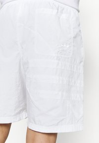 Bílé sportovní šortky z lehkého materiálu, s přišitou kapsou, horizontálním panelem a decentním logem na boku.