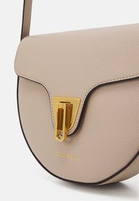 Borsa a tracolla in pelle beige con forma curva, hardware dorato e una chiusura distintiva. Presenta un logo impresso e cuciture a contrasto.