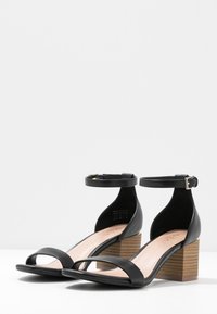 Sandales en cuir noir avec une large bride à l'avant, une bride à la cheville et un talon bloc en bois empilé. Texture lisse et design ouvert.