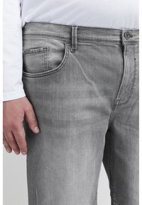 Graue Jeans aus Denim mit klassischem Schnitt, ausgestattet mit einer Fronttasche, sichtbaren Nähten und einer glatten Textur. Getragen mit einem weißen Langarmhemd.