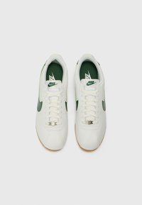 Tênis brancos com detalhes em verde, apresentando um cabedal de couro liso, bico redondo e uma sola interior texturizada em verde. Design com atacadores e sola de borracha natural.
