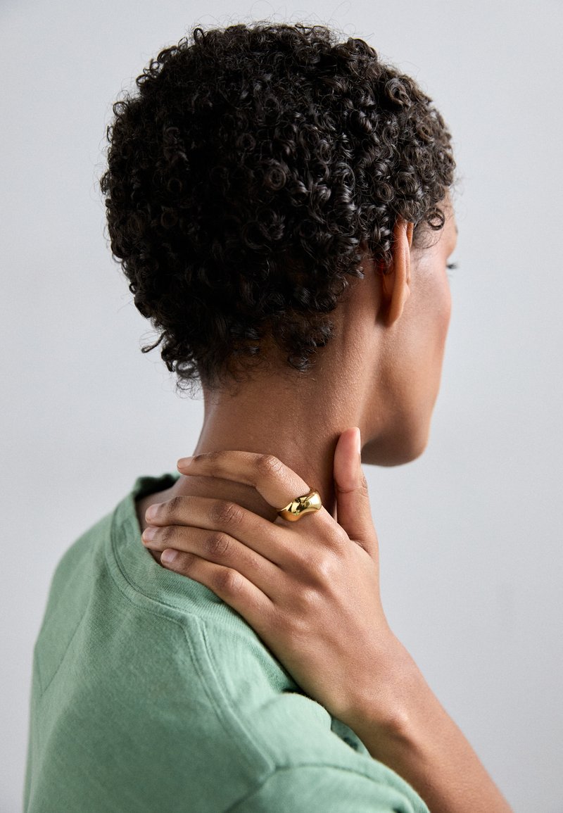 Goldring mit einer glatten, polierten Oberfläche, getragen an einer Hand, die auf einem Hals ruht. Das Modell trägt ein grünes, strukturiertes T-Shirt mit einem runden Ausschnitt.