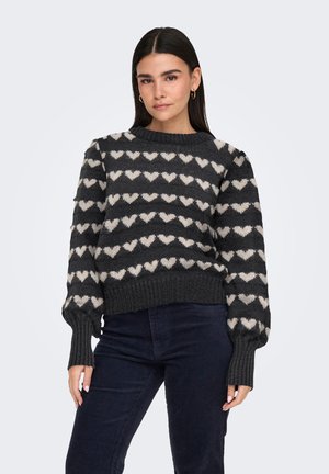 Kvinde med langt mørkt hår iført en sort sweater med hvide hjerte-mønstre og mørke bukser mod en ensartet lys baggrund.