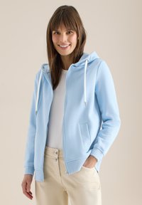 Femme souriante portant un sweat à capuche bleu clair zippé sur un t-shirt blanc, pantalon beige, debout devant un fond uni avec une main dans la poche.