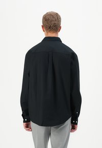 Man met kort krullend haar, gekleed in een losse zwarte longsleeve en lichtgrijze broek, met zijn rug naar de camera gericht tegen een eenvoudige witte achtergrond.