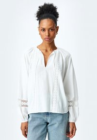 Hvid bomuldsbluse med V-hals, blonde detaljer langs fronten og ærmerne, samt sammensnørrede skuldersømme. Bæres med lyseblå jeans.