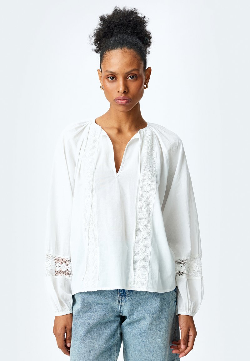 Hvid bomuldsbluse med V-hals, blonde detaljer langs fronten og ærmerne, samt sammensnørrede skuldersømme. Bæres med lyseblå jeans.