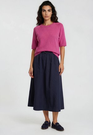 Woman with dark hair wearing a pink short-sleeve sweater, navy blue midi skirt, and navy blue loafers standing against a plain white background.
