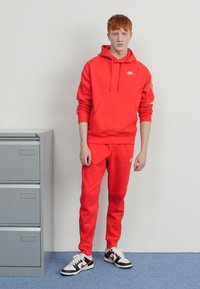 Rdeča Nike kapuca in hlače iz mehke tkanine, z sprednjim žepom, logotipnimi detajli in v kombinaciji s črnimi in belimi športnimi čevlji. Raven ozadje.