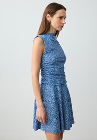 Blauwe jurk met witte stippen, mouwloos ontwerp, hoge halslijn, aansluitende lijfje en uitlopende rok. Zachte stof met een gladde textuur.