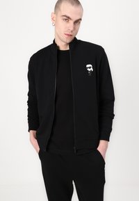 Black zip-up jacket made of smooth fabric. Features a cartoon logo on the left chest, high collar, and fitted cuffs. Paired with black pants.