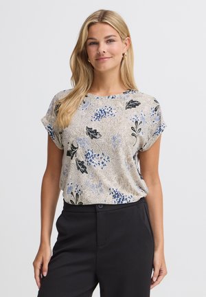 Femme aux longs cheveux blonds portant un chemisier beige à manches courtes fleuri et un pantalon noir, souriant devant un fond clair uni.