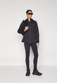 Juodas zip-up striukė su kapišonu ir logotipu, derinama su tamsiomis kelnių tipo legging'ais su šoninėmis juostelėmis ir juodais žygio batais. Papildomi aksesuarai – saulės akiniai.