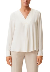 Blouse blanche à manches longues avec un col en V, tissu doux, coupe décontractée et plis subtils à l'avant. Portée avec un pantalon beige clair.