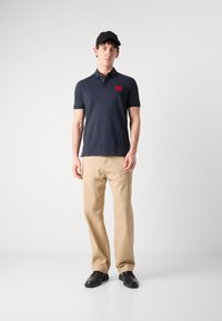 Marineblå polo med en rød logo-patch, beige bukser og sorte sko. Skjorten har klassisk krave og korte ærmer.