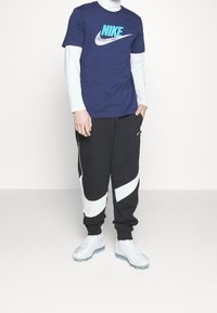 Marinblå Nike t-shirt med vit logotyp, lager på lager över en vit långärmad tröja. Svarta joggarbyxor med vitt virvelfmönster, parat med vita sneakers.