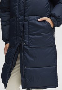 Navy blue puffer coat with a zip closure, side pockets, and quilted texture. Features elastic cuffs and contouring seams for shape.
