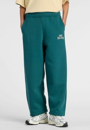 Personne portant un pantalon New Balance ample de couleur bleu sarcelle et des baskets blanches New Balance, debout les mains dans les poches.