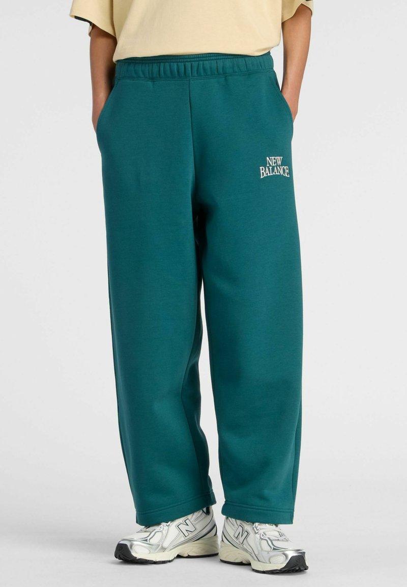 Personne portant un pantalon New Balance ample de couleur bleu sarcelle et des baskets blanches New Balance, debout les mains dans les poches.