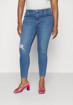 Kvinde iført blå slidte skinny jeans og lyserøde høje hæle med stropper, stående mod en ensfarvet lys baggrund.