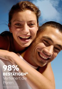 Zwei lächelnde Personen mit heller Haut, eine mit dunklen Haaren und einem weißen Shirt, die andere mit braunen Haaren, vor einem blauen Himmel.