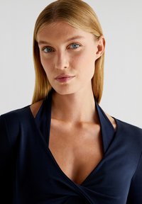 Navyblauw top met een diepe V-hals, zachte stof en een gedraaide detail bij de buste, met halter nek en aansluitende lange mouwen.