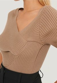 Ribbad beige topp med V-ringning, med en texturerad mönster och böjda sömmar. Kombinerad med svart underdel som visar en tajt silhuett.