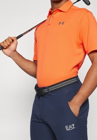 Oranžový polo tričko s krátkým rukávem a texturovaným vzorem, kombinované s námořnicky modrými kalhotami s páskem se pruhovaným designem a logem na kapse.