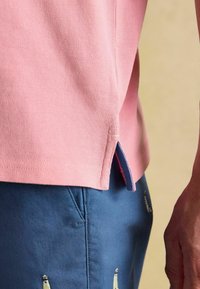 Gaiši rozā polo krekls ar teksturētu audumu un sānu ventilācijas atverēm, apvienots ar zilām biksēm, kurām ir izšūti raksti. Tuvplāns detaļām.