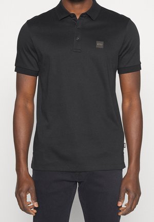 Man wearing a fitted black short-sleeve polo shirt with a small square logo patch on the left chest.