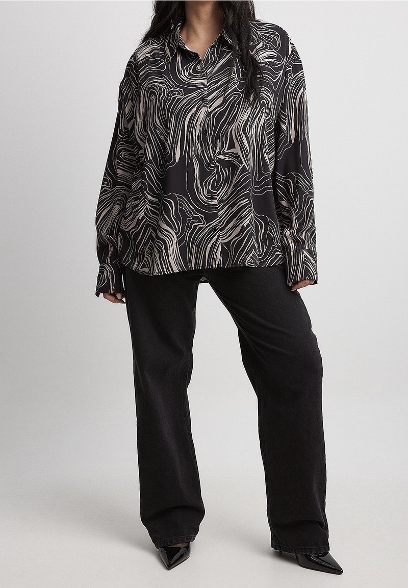 Blusa nera a maniche lunghe con un motivo ondulato crema e beige, realizzata in tessuto morbido, abbinata a pantaloni neri a gamba larga e tacchi a punta.