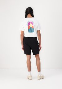 Tricou alb cu un grafic colorat al munților și textul "Columbia Sportswear Company", asortat cu pantaloni scurți negri și adidași bej.