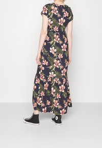 Robe maxi fleurie à manches courtes, fond bleu marine, motif floral rose et vert, design à volants, associée à des baskets montantes noires.