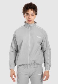 Giacca zip grigia chiara realizzata in materiale leggero e liscio, con piping bianco lungo le spalle, abbinata a pantaloni grigi coordinati.