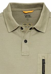 Beige Poloshirt mit einem gerippten Kragen, zwei schwarzen Knöpfen und einer mit Reißverschluss versehenen Brusttasche. Der Stoff wirkt texturiert und hat ein atmungsaktives Design.