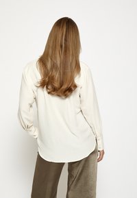 Kreemivärviline, nööpidega pluus pika varrukaga, lõdva lõikega, tagantvaade, mis näitab tekstureeritud kangast. Koos õlavarre pealt lõdva sobitumisega oliivrohelised püksid.