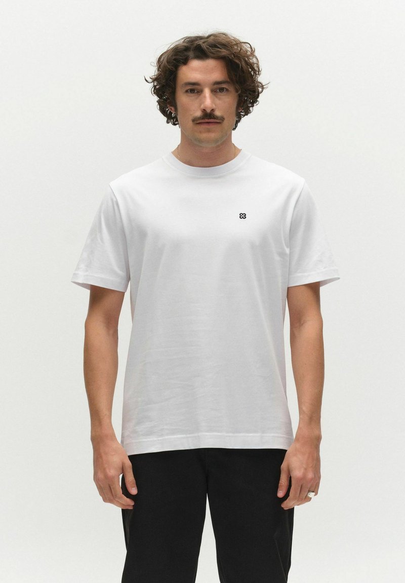 Homme aux cheveux bouclés et à la moustache portant un t-shirt blanc uni à manches courtes avec un petit logo noir sur la poitrine et un pantalon noir.