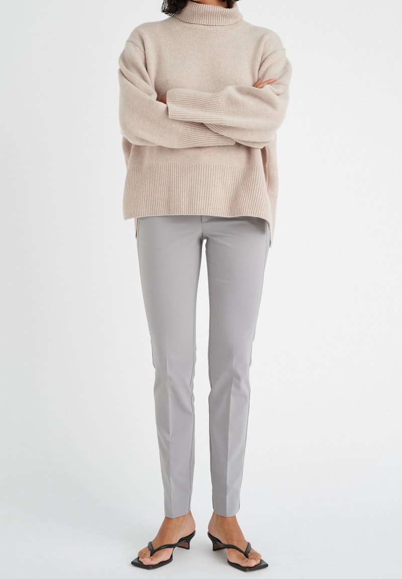 Béžový pletený svetr s vysokým límcem; volný střih. Světle šedé kalhoty s úzkým střihem. Černé sandály na podpatku. Jednoduché pozadí.