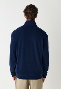 Námořnická flísová bunda s límcem, volným střihem, dlouhými rukávy a rovný lem. Hladká textura s čistým, minimalistickým designem.