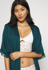 Quimono de cetim verde-água com rendas nas mangas, usado sobre um bralette de renda branca e roupa interior combinando, com um design em forma de olho-chave.